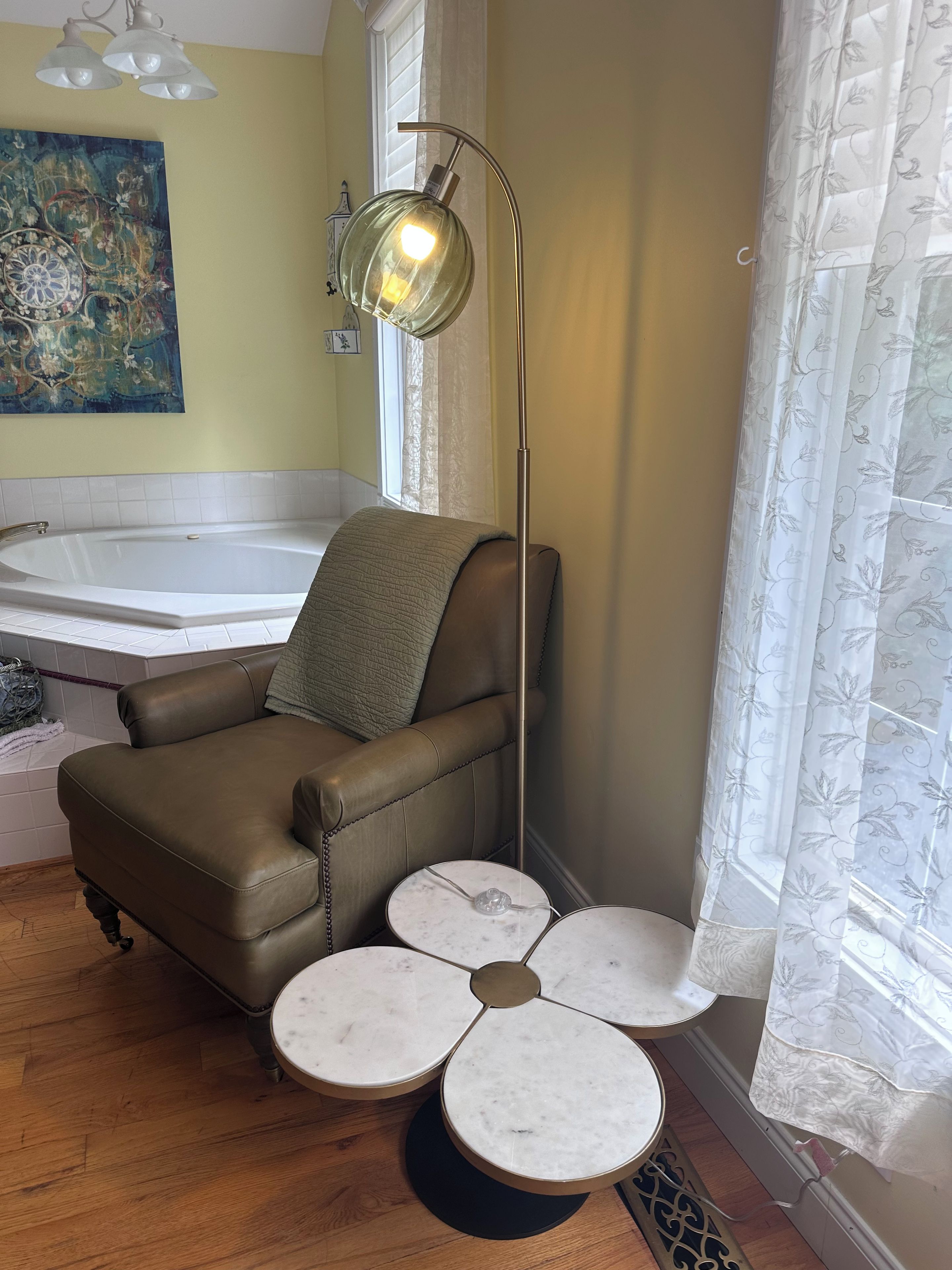 Une salle de bain avec une baignoire, un fauteuil en cuir marron, une lampe sur pied et une table ronde à quatre pieds.