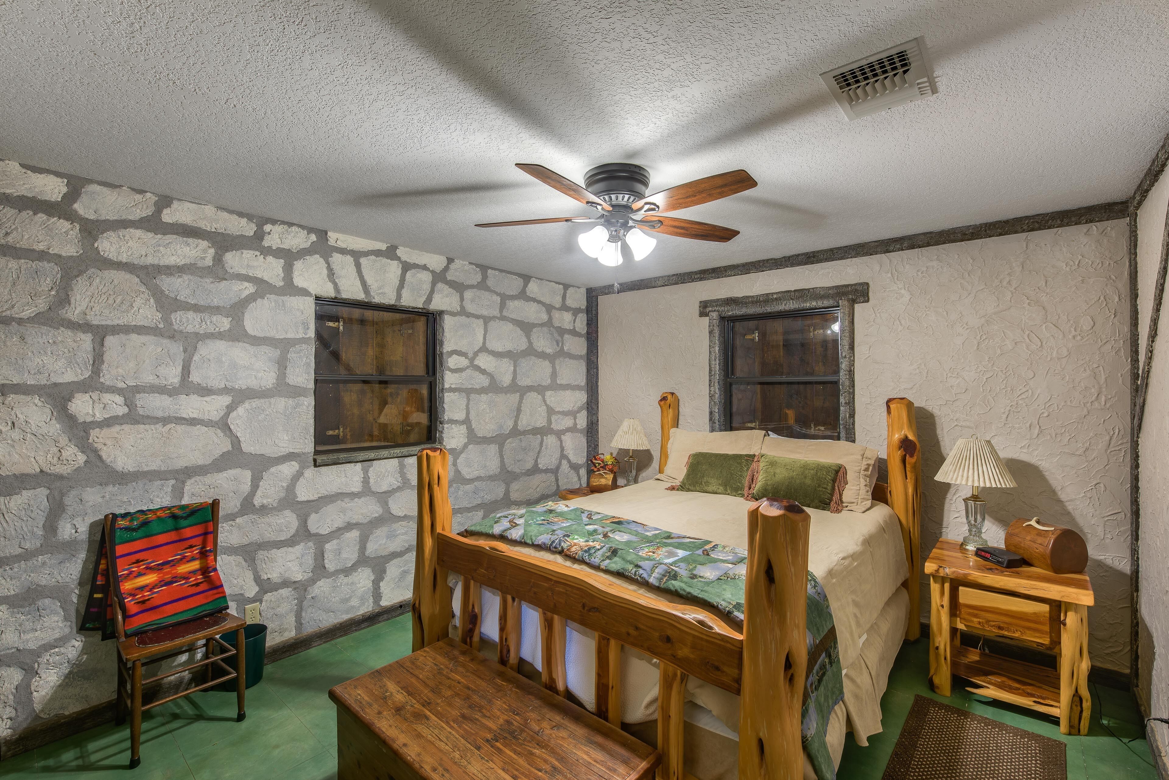 A bedroom with a stone wall, a wooden bed, a chair, a bench, and a ceiling fan.
