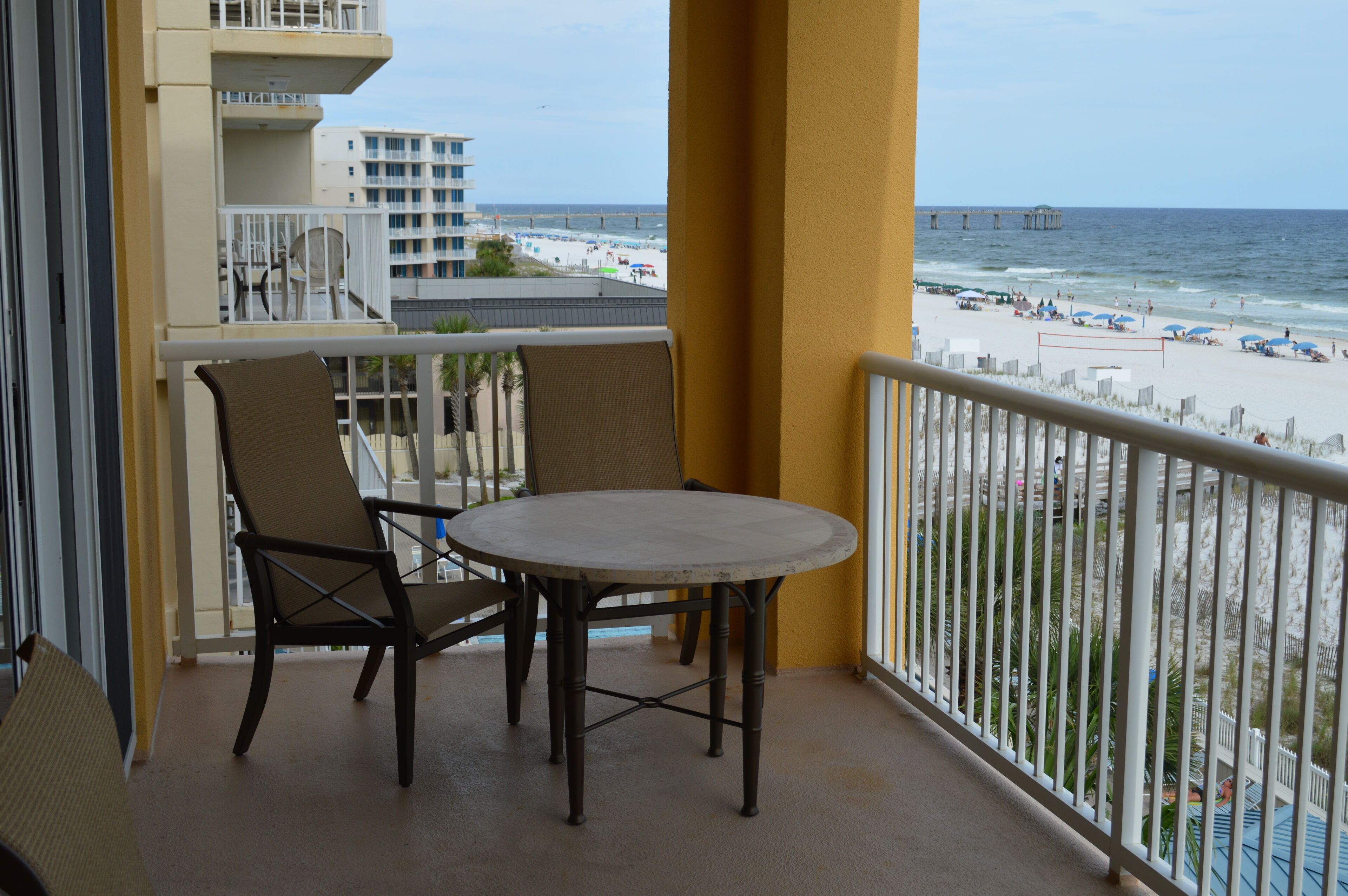 A balcony with a table and chairs, overlooking a beach with umbrellas and a pier.