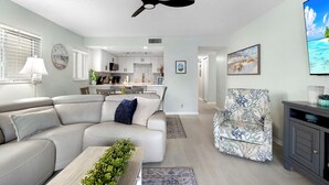Una sala de estar moderna con un sofá seccional, un sillón estampado, un ventilador de techo y una cocina al fondo.