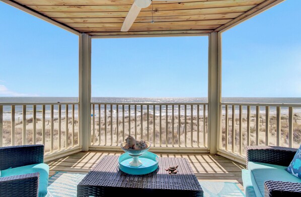 Área externa coberta com vista para a praia, mobiliada com cadeiras de vime e uma mesa.