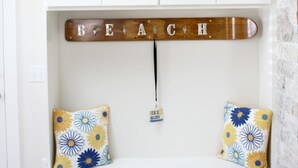 Un banco blanco con cojines estampados con flores, un letrero de madera que dice 'BEACH' y una pequeña etiqueta colgando del letrero.