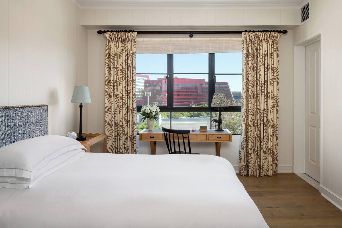 A hotel room with a bed, a desk with a chair, a lamp, and a window with curtains.