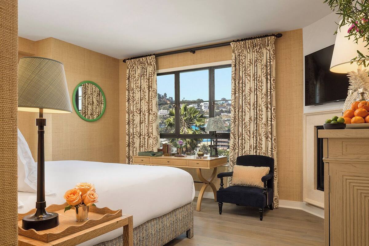 A hotel room with a bed, desk, chair, and a view of a cityscape through a window.