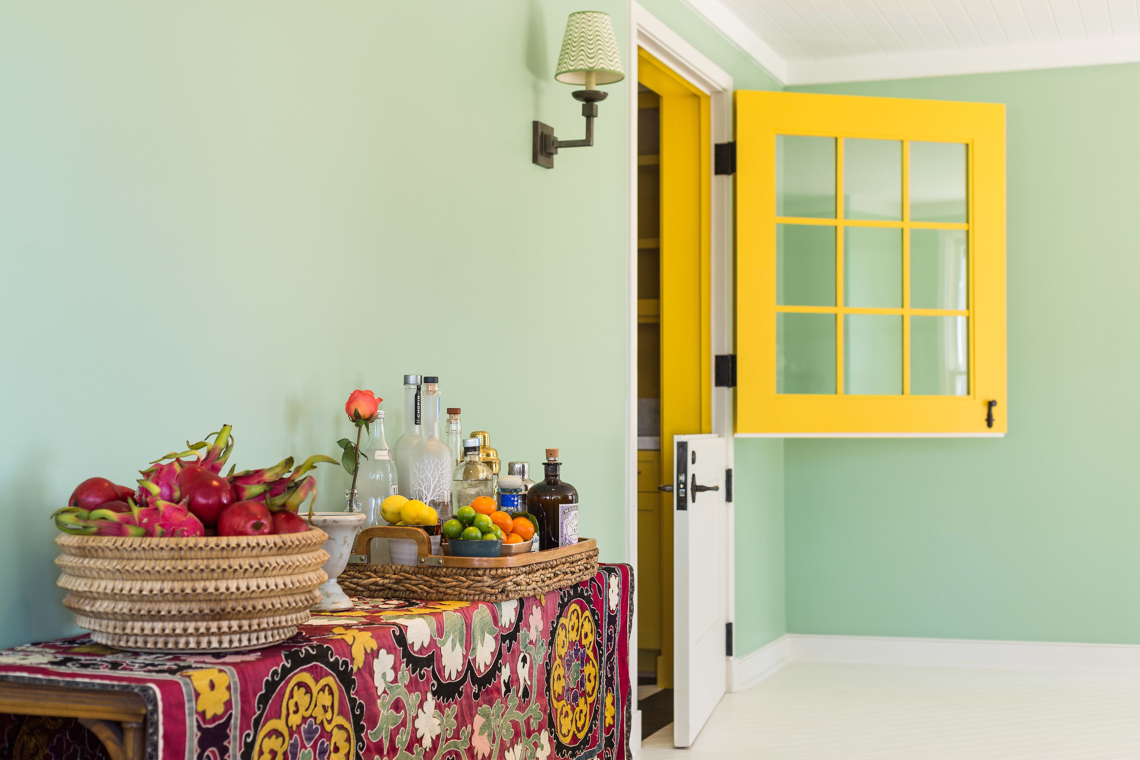 Une pièce avec une table dressée, ornée de fruits et de bouteilles, une porte jaune et une lampe fixée au mur.