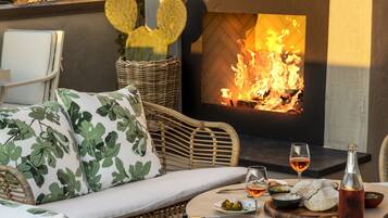 A cozy outdoor seating area with wicker furniture, a table with a bottle and glasses, and a fireplace.