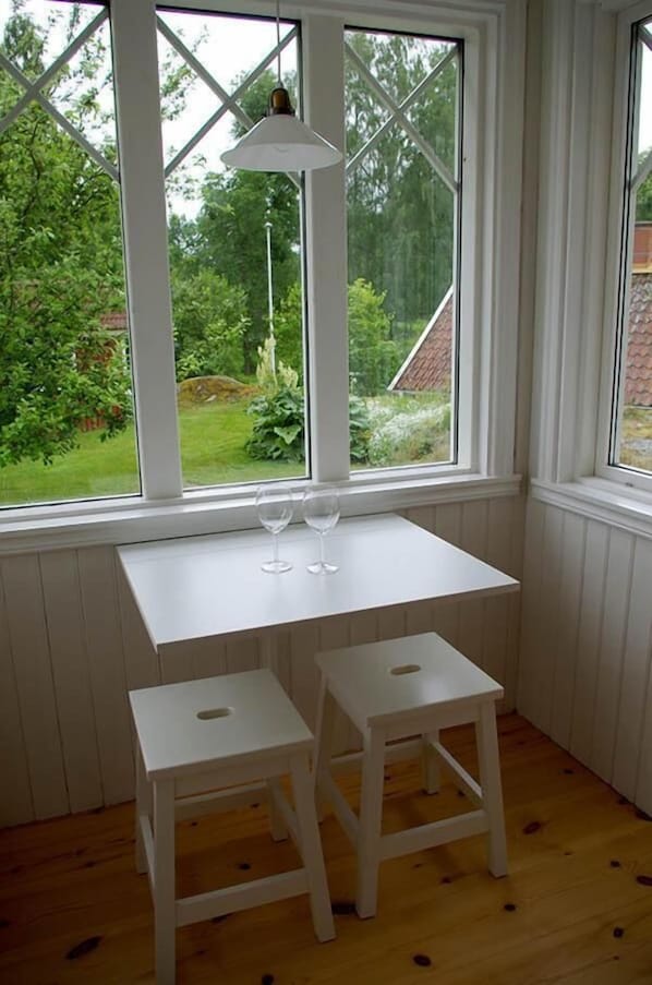 Habitación de esquina con una mesa blanca y dos taburetes, una ventana con vistas a zonas verdes, y una lámpara colgante.