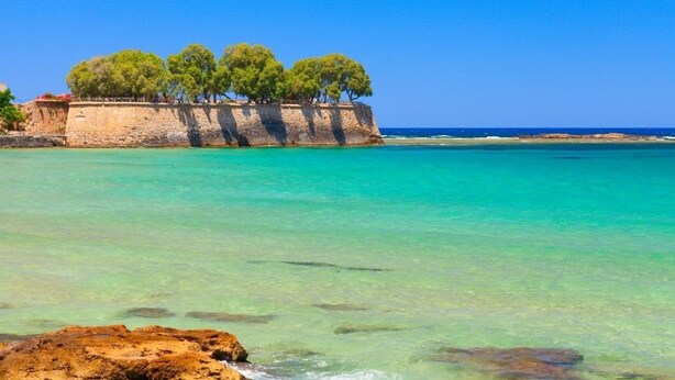 A coastal scene with clear turquoise waters, a rocky shoreline, and a stone wall with trees. - Ranias luxurious Apartment with sea view! (Chania)