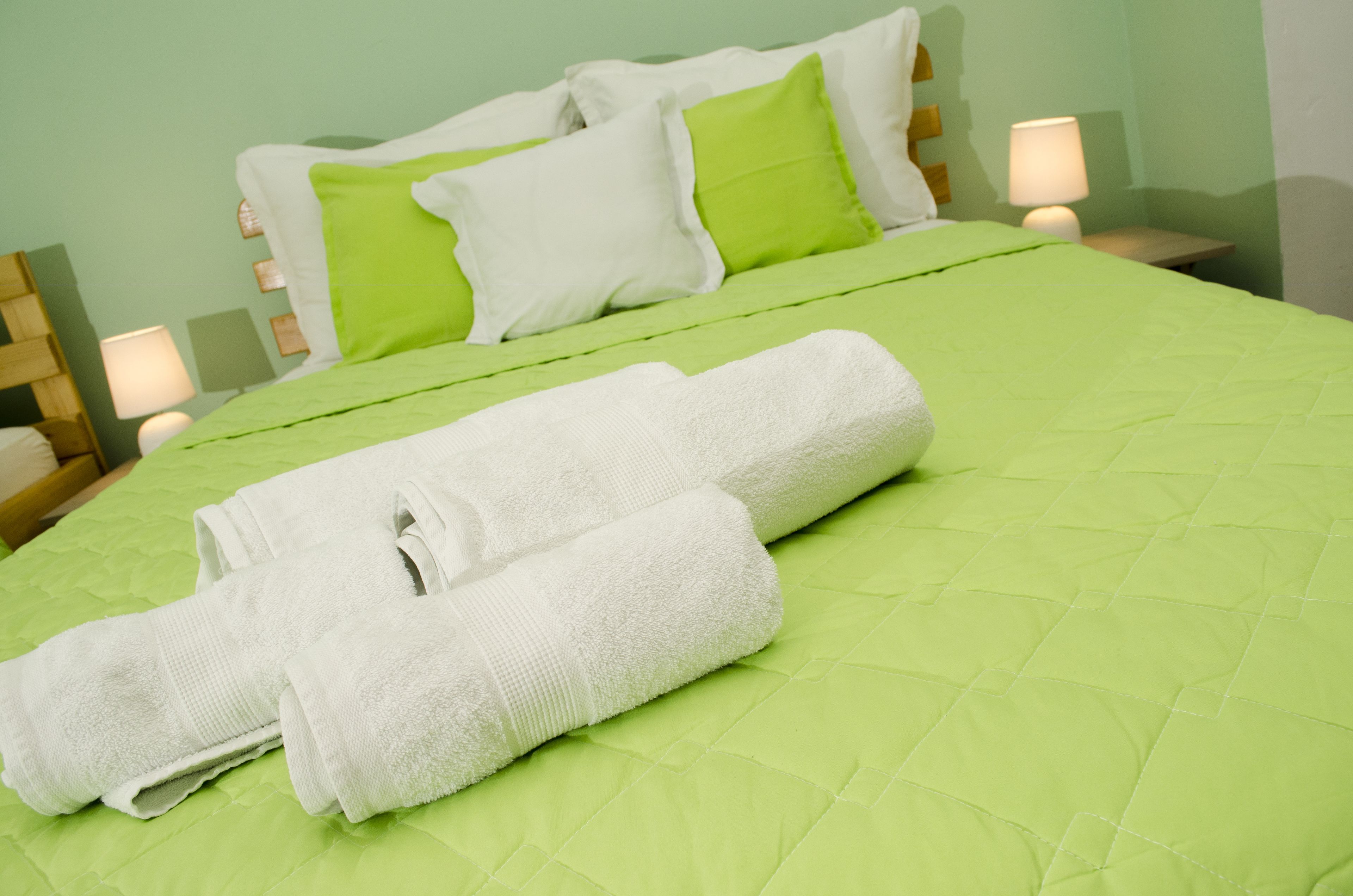 Una cama bien hecha con colcha verde, almohadas blancas y toallas enrolladas en blanco.