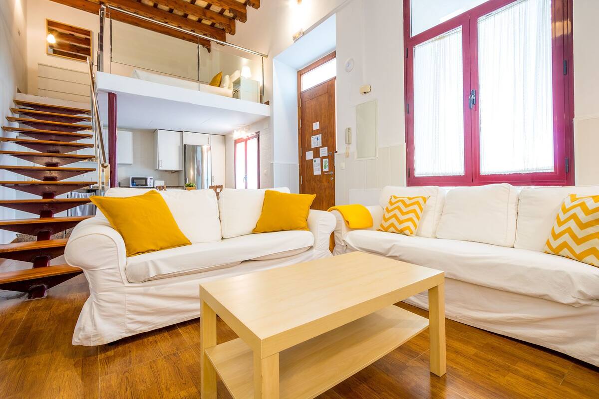 Una sala de estar moderna con un sofá blanco, una mesa de centro de madera y una escalera que sube a un nivel superior.