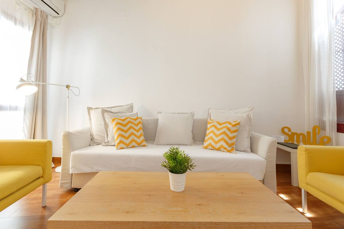 Una sala de estar moderna con un sofá, dos sillones amarillos, una mesa de centro con una planta y una lámpara de pie.