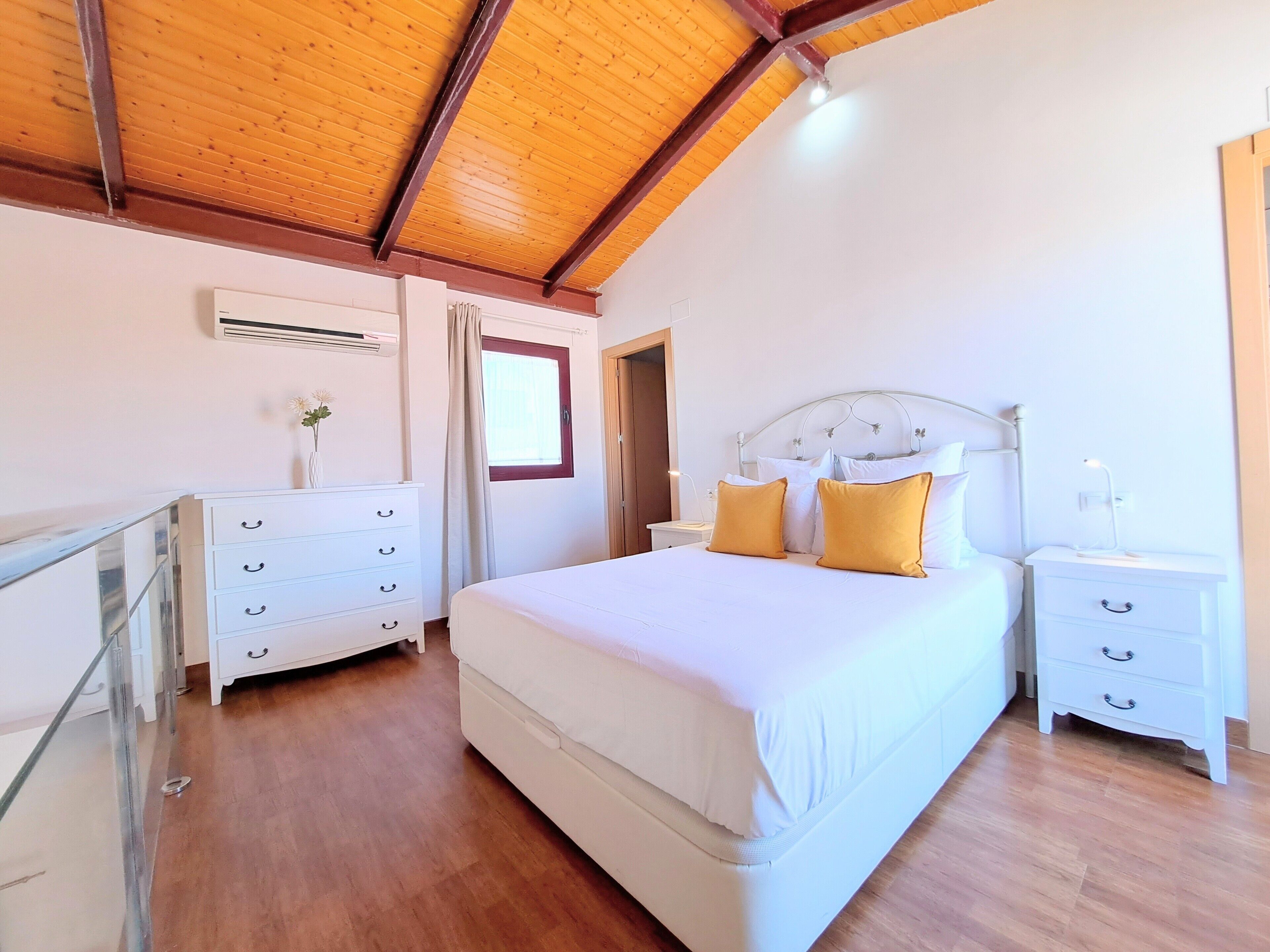 Un dormitorio con techo de madera, una cama blanca con almohadas amarillas, un tocador blanco y una mesita de noche con una lámpara.