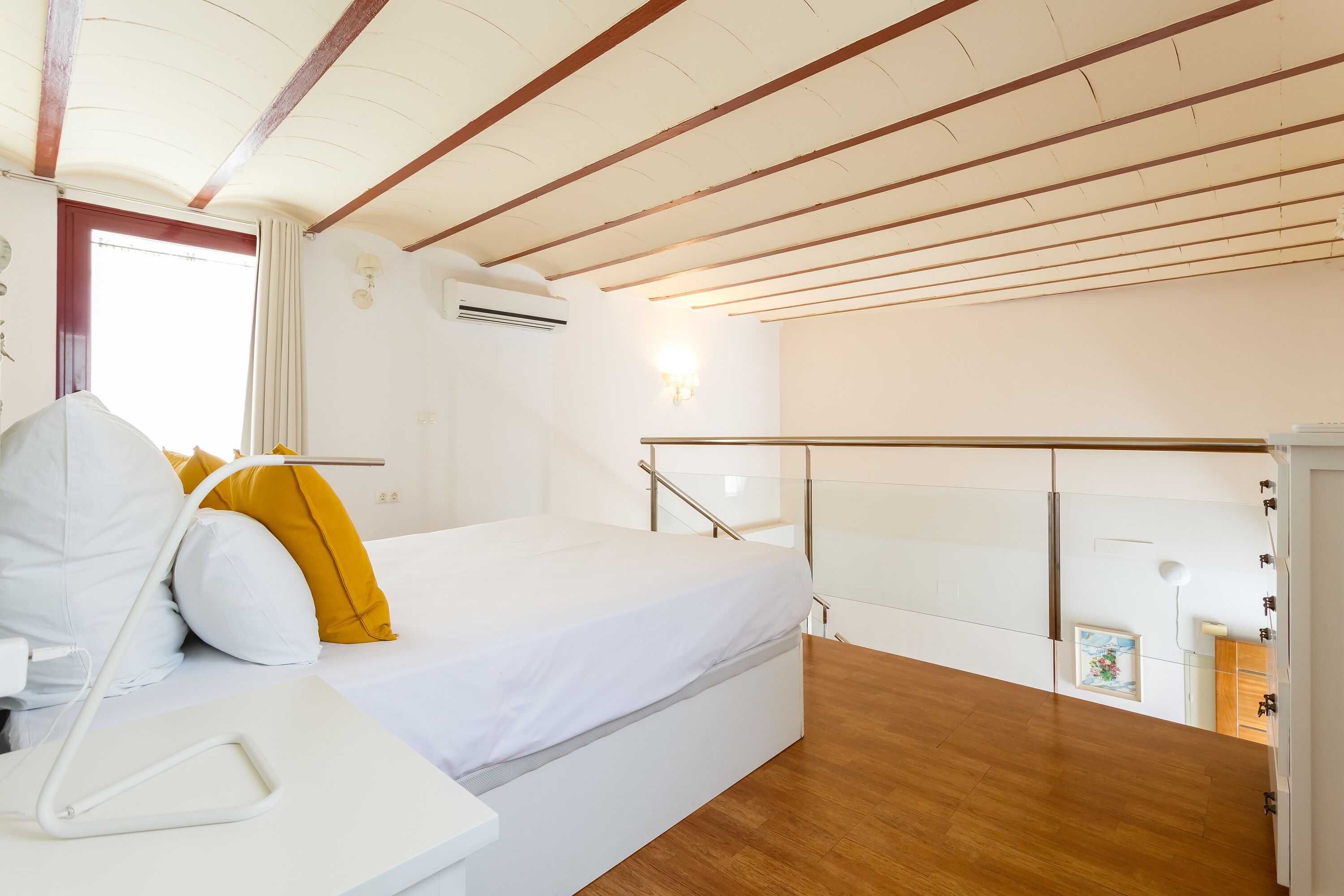 Un dormitorio con cama, mesita de noche, ventana con cortinas, suelo de madera y barandilla de cristal.