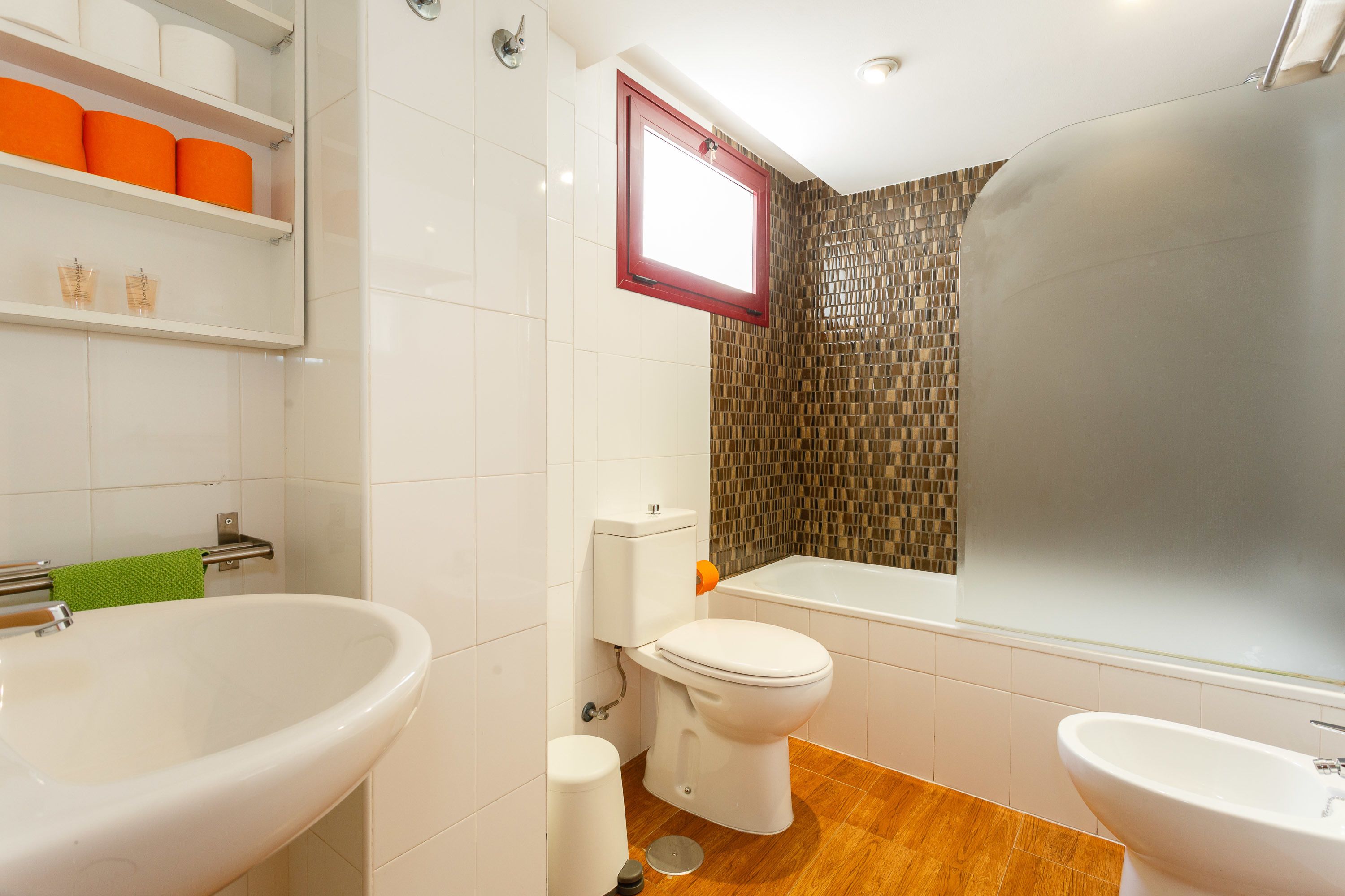 Une salle de bain avec un grand lavabo blanc, des toilettes et une baignoire. On y trouve des étagères avec des articles de toilette et une fenêtre au-dessus des toilettes.