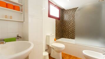Une salle de bain avec un grand lavabo blanc, des toilettes et une baignoire. On y trouve des étagères avec des articles de toilette et une fenêtre au-dessus des toilettes.