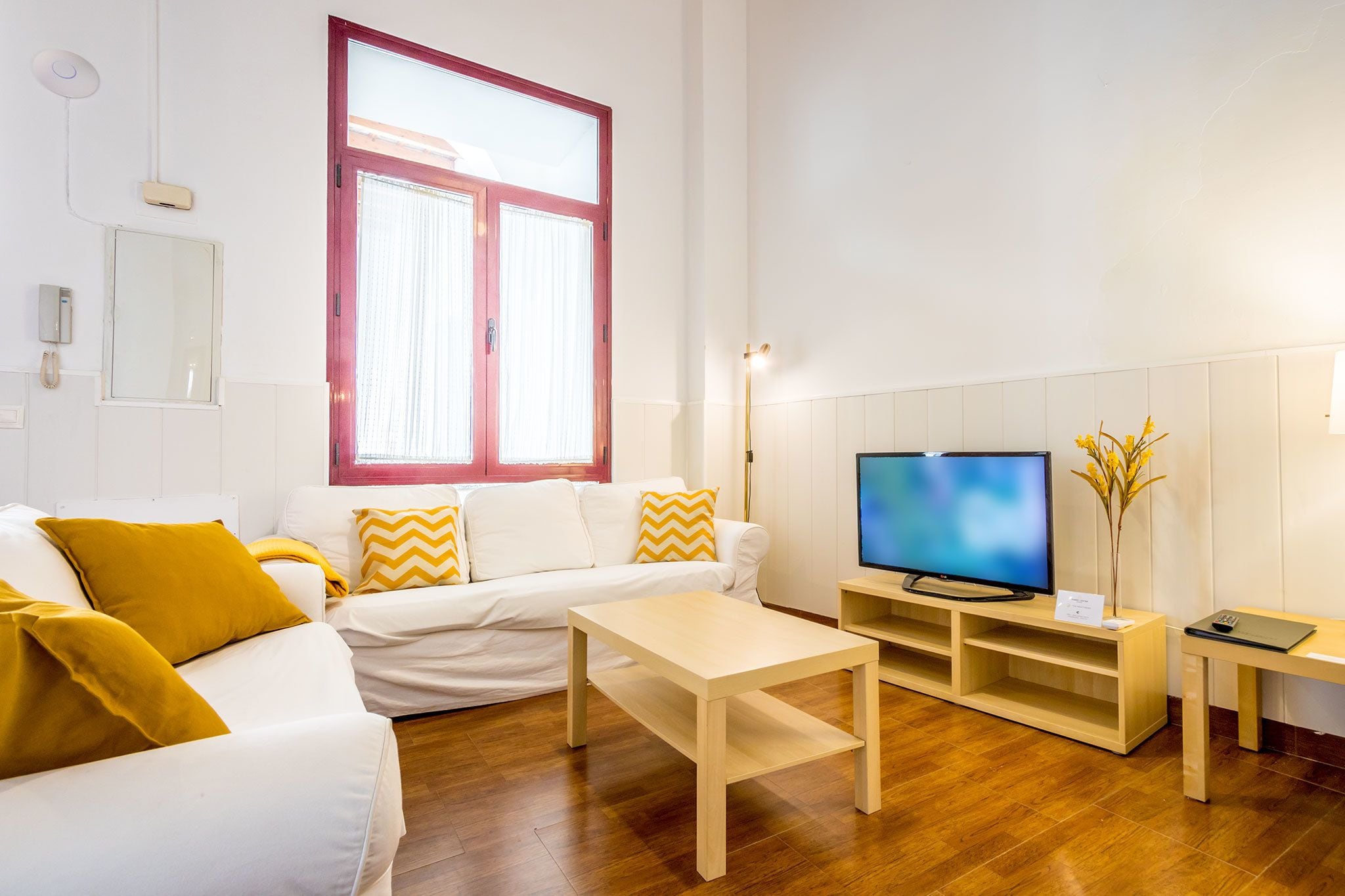 A modern living room with a white sofa, yellow pillows, a wooden coffee table, a TV on a wooden stand, and a large window with curtains.