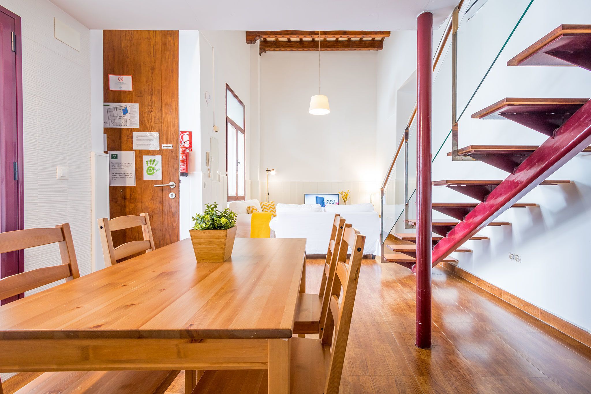 A dining area with a wooden table and chairs, a potted plant, and a staircase with red railings.