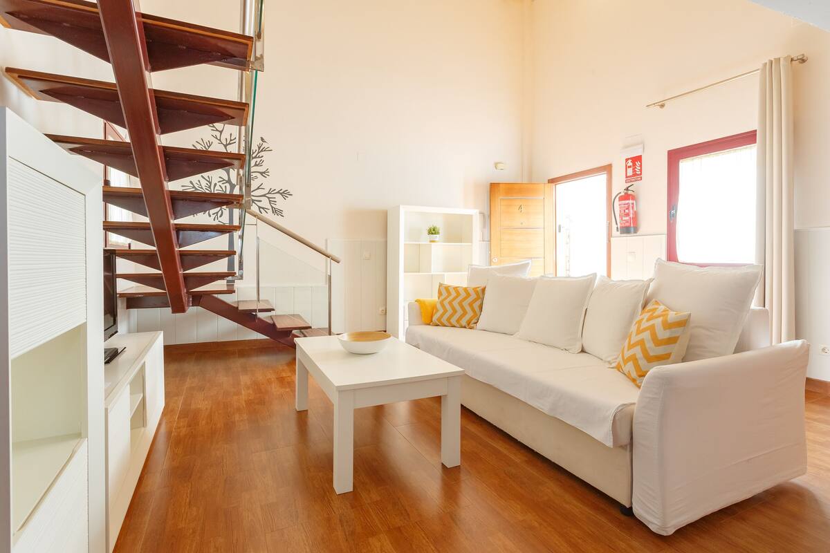 Una sala de estar moderna con una escalera de madera, un sofá blanco y una pequeña mesa blanca.
