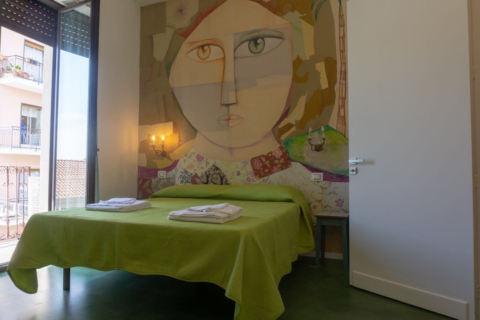 A hotel room with a bed, a green bedspread, a wall mural, and a balcony with a railing.