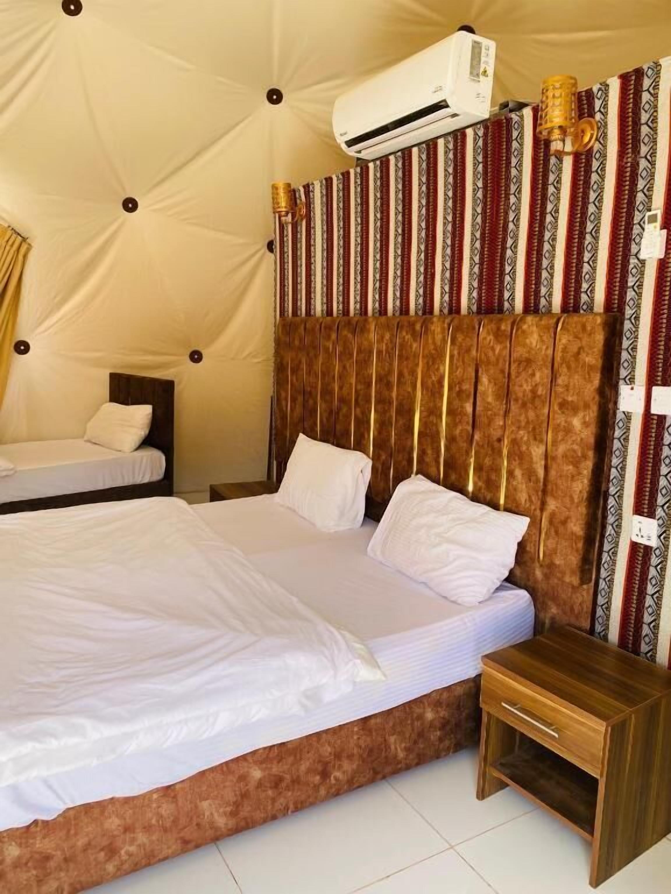 A bedroom with two beds, a wooden nightstand, a patterned curtain, and an air conditioning unit.