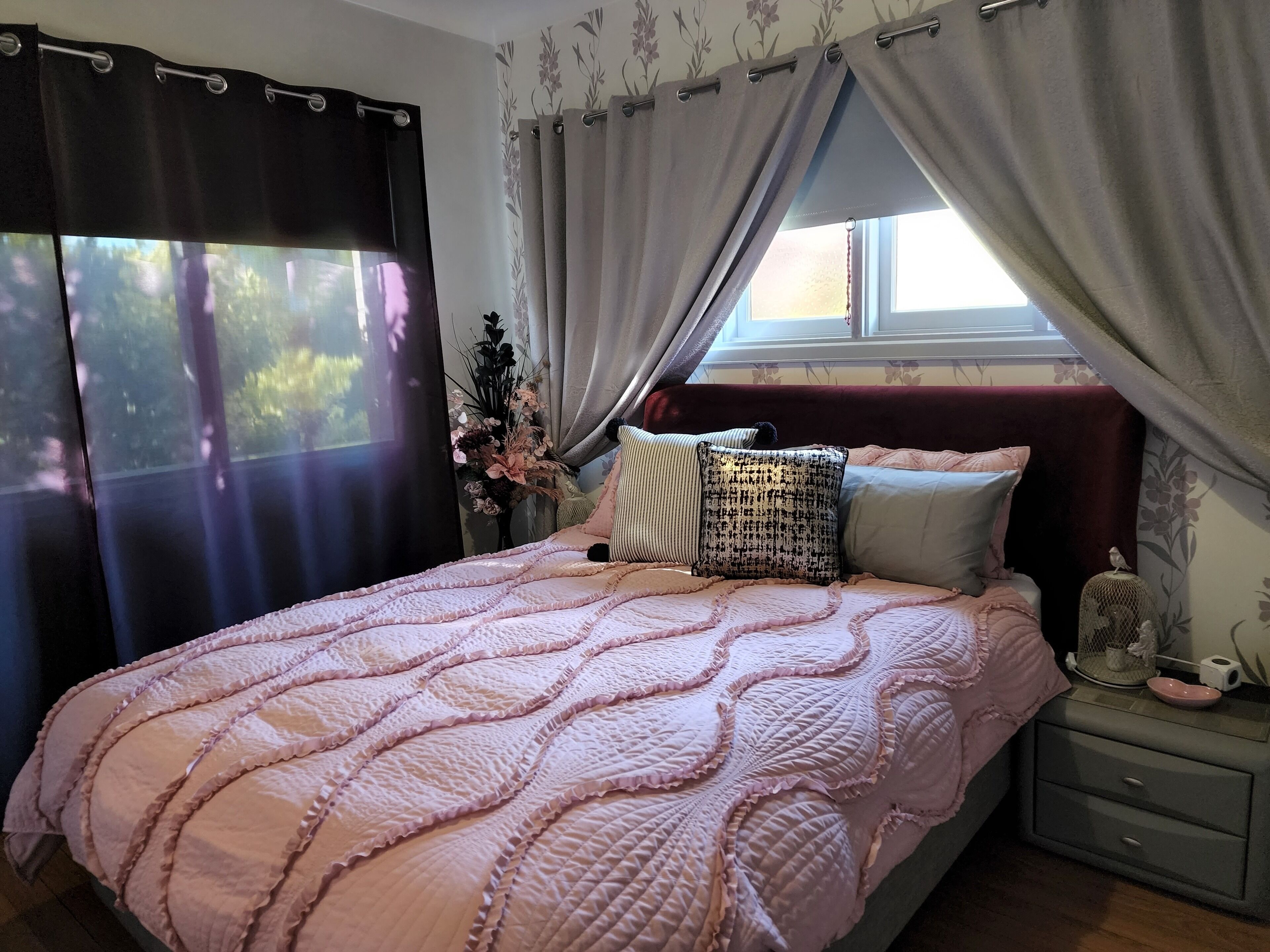 A bedroom with a bed, a nightstand, a window with curtains, and a floral wall pattern.