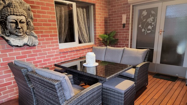 Un patio con mobiliario de mimbre, una mesa de vidrio y una escultura de cabeza de Buda sobre un muro de ladrillo.