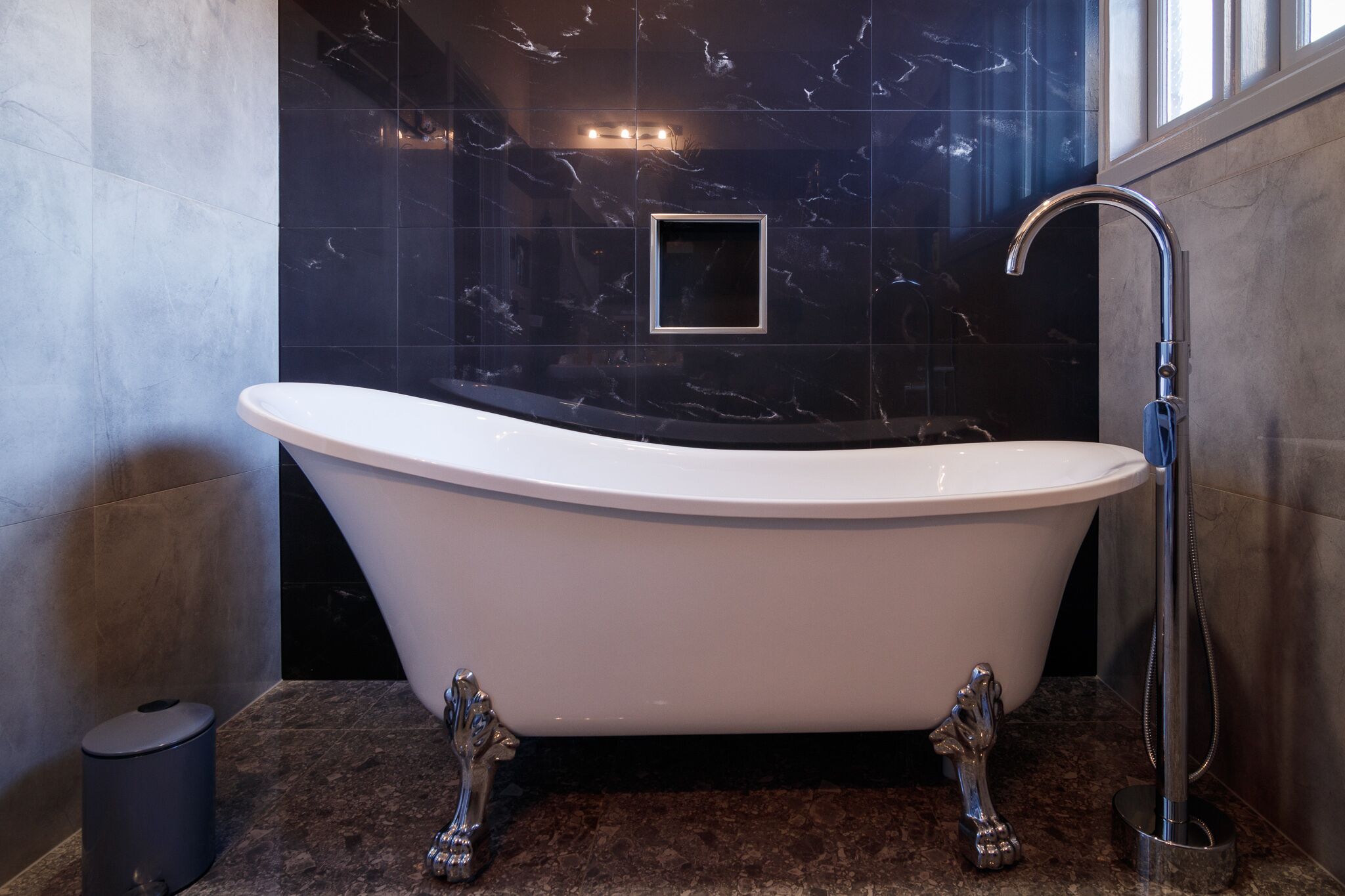 Une baignoire sur pied, dotée d’un robinet moderne, est placée devant un mur de marbre foncé, agrémenté d’un petit miroir carré.