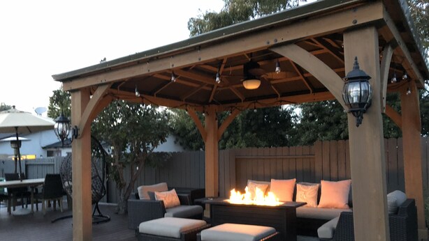 Een terras buiten met een houten pergola, rieten meubels, een vuurkorf en een eethoek met een parasol.