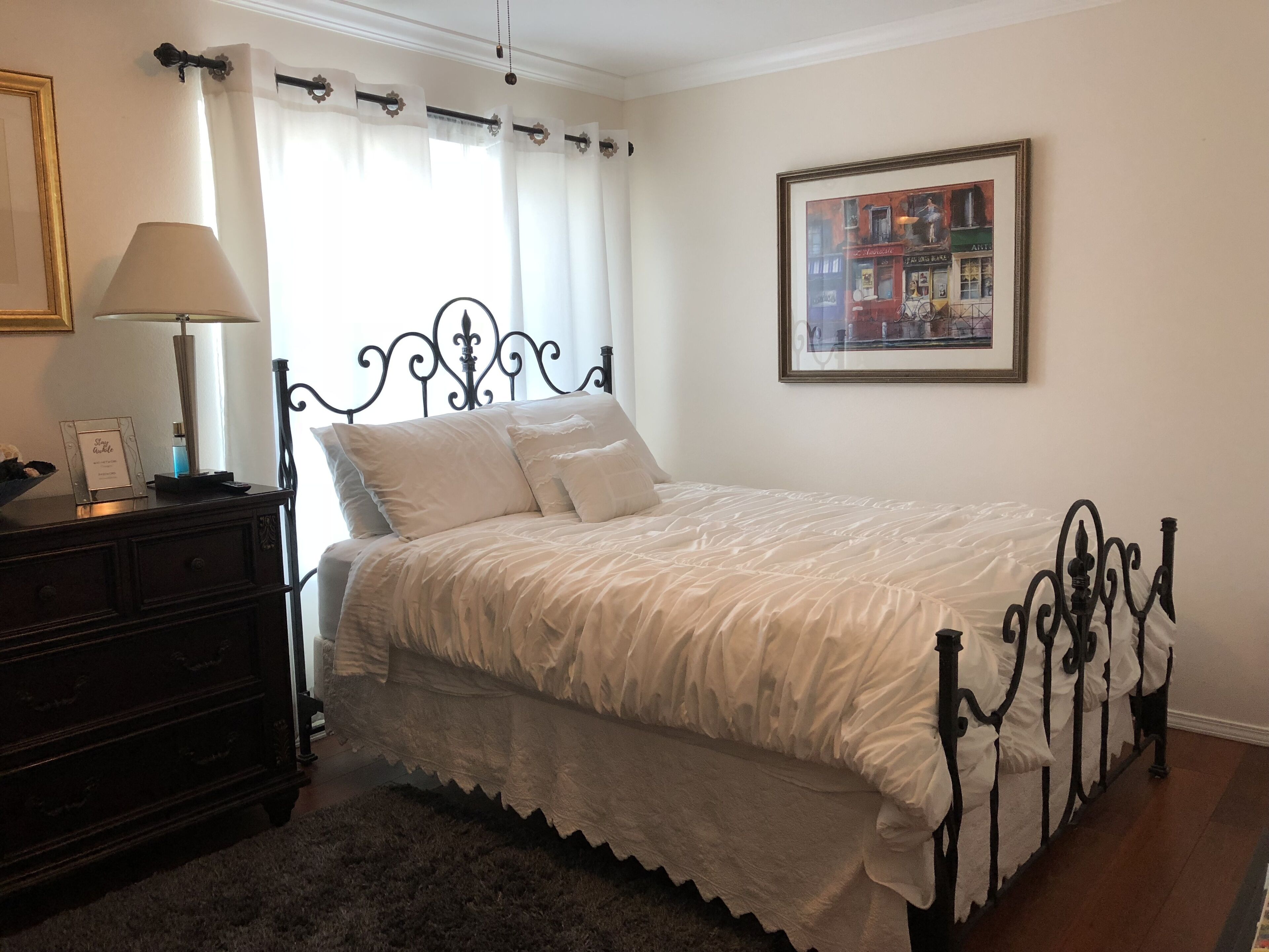 A neatly made bed with a white bedspread and pillows, a dark wooden dresser with a lamp, and a framed picture on the wall.
