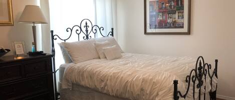 A neatly made bed with a white bedspread and pillows, a dark wooden dresser with a lamp, and a framed picture on the wall.