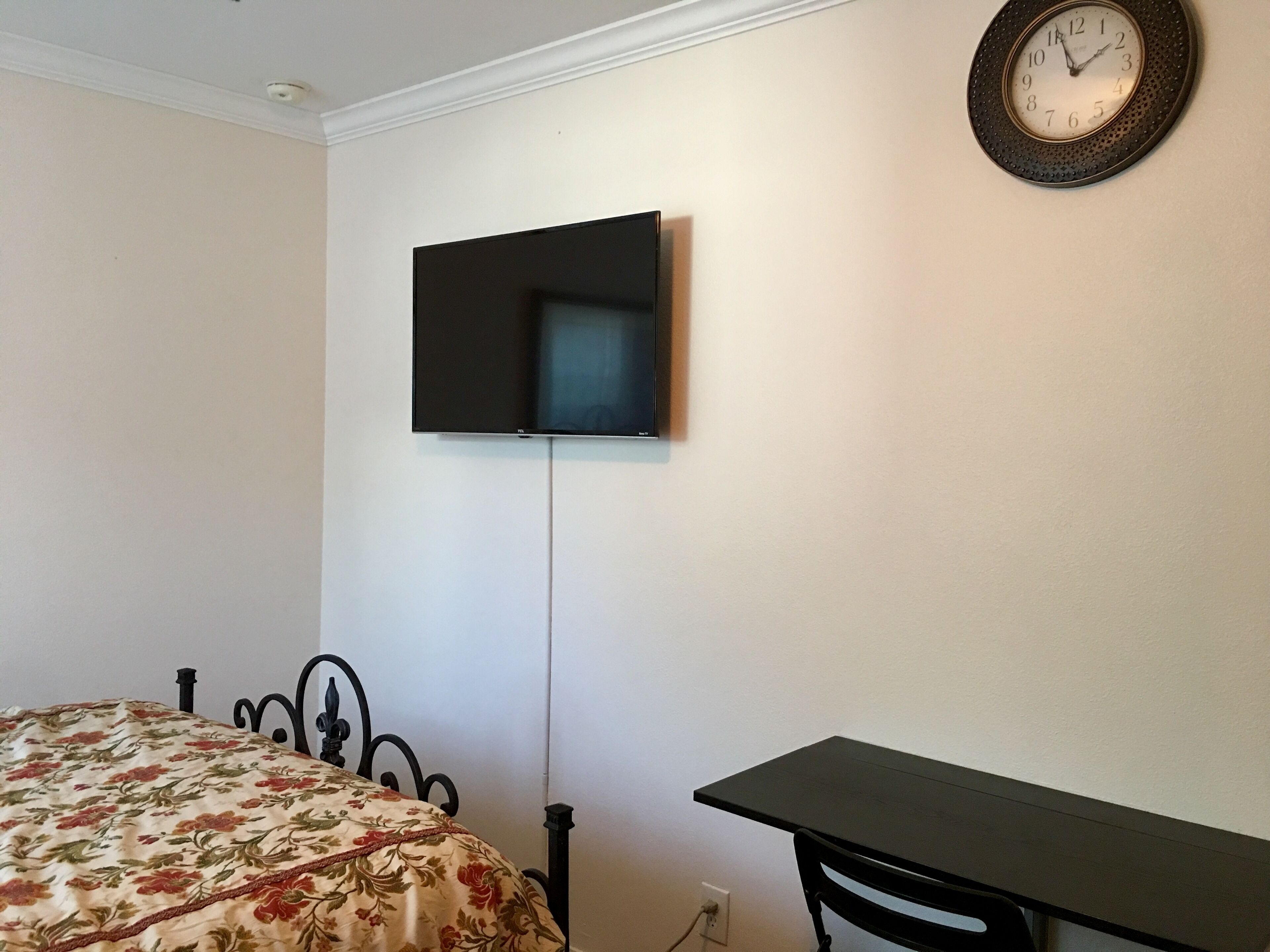 Quarto com cama, uma mesinha, televisão na parede e um relógio.