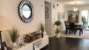 Een moderne woonkamer met een donker houten vloer, een witte console met wijnflessen, een grote spiegel en een eethoek met tafel en stoelen.