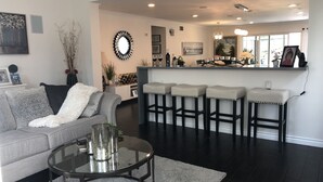 Een moderne woonkamer met een grijze bank, een glazen salontafel en een bar met krukken.