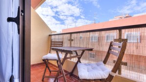 En balkon med et træbord og to stole, et flettet hegn og udsigt til bygninger under en klar himmel.