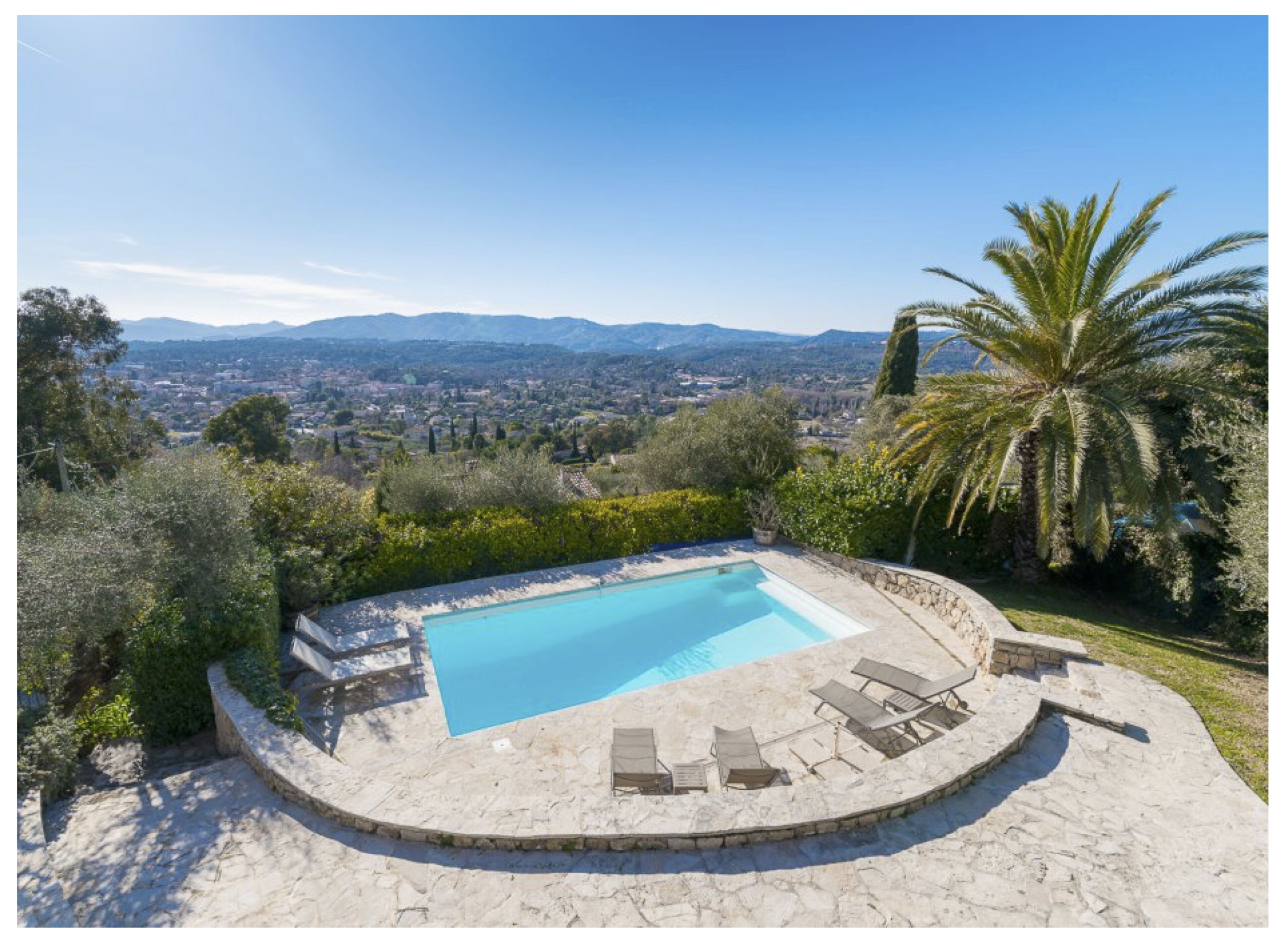 A swimming pool with a stone deck and lounge chairs, surrounded by greenery and palm trees, overlooking a cityscape.