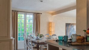 Een eethoek met een tafel gedekt voor vier personen, witte stoelen, een houten bank, een raam met gordijnen en een schilderij aan de muur.