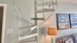 Un escalier en colimaçon avec une rampe en métal blanc, une lampe sur une table d’appoint et des coussins à rayures sur un canapé.