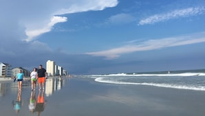 Une plage avec un ciel dégagé, quelques nuages et des bâtiments en arrière-plan.