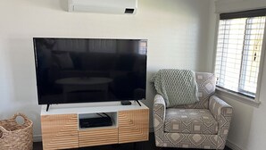 A living room with a flat-screen TV on a wooden TV stand, a patterned armchair, a large floral rug, and a window with blinds.