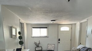 A living room with a wooden floor, a grey sofa, a dining table with chairs, a white cabinet, a rug, and a window with blinds.