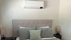 A bedroom with a bed, two bedside tables with lamps, a wall-mounted air conditioner, and a nightstand with a power strip.