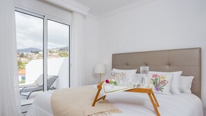 A hotel room with a large bed, a bedside table with a lamp, and a view of the city through a large window.