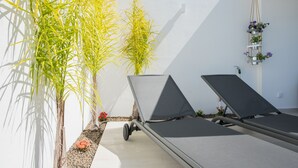 Un espace extérieur ensoleillé avec deux chaises longues, un petit pot de fleurs orné de fleurs rouges et de grandes plantes vertes adossées à un mur blanc.