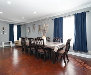 A dining room with a wooden table and chairs, blue curtains, and a vase with branches.