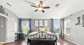 A bedroom with a bed, a ceiling fan, a window with curtains, and a wall with framed text.