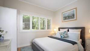 A bedroom with a bed, bedside tables, a nightstand, a window with blinds, and a painting on the wall. - Dogs Stay Free. Cosy Restful Retreat with Fireplace. (Wentworth Falls)