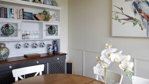 A dining area with a wooden table, white chairs, a bookshelf with books and decorative items, a stained glass window, and a framed painting of a bird on a branch. - Dogs Stay Free. Cosy Restful Retreat with Fireplace. (Wentworth Falls)