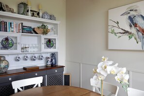 A dining area with a wooden table, white chairs, a bookshelf with books and decorative items, a stained glass window, and a framed painting of a bird on a branch. - Dogs Stay Free. Cosy Restful Retreat with Fireplace. (Wentworth Falls)