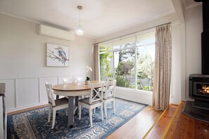 A dining area with a round table, white chairs, a blue patterned rug, a painting of a bird, and a fireplace. - Camellia Cottage. Where Dogs Holiday Too. (Wentworth Falls)