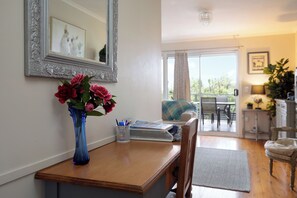 A room with a wooden desk, a vase with flowers, a chair, a dining area with a table and chairs, and a view through a glass door. - Dogs Stay Free. Cosy Restful Retreat with Fireplace. (Wentworth Falls)
